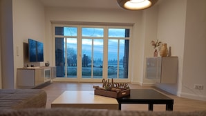 Una sala moderna con un gran ventanal corredera que da a un área exterior nevada, un televisor de pantalla plana sobre un mueble bajo y una mesa de centro minimalista.