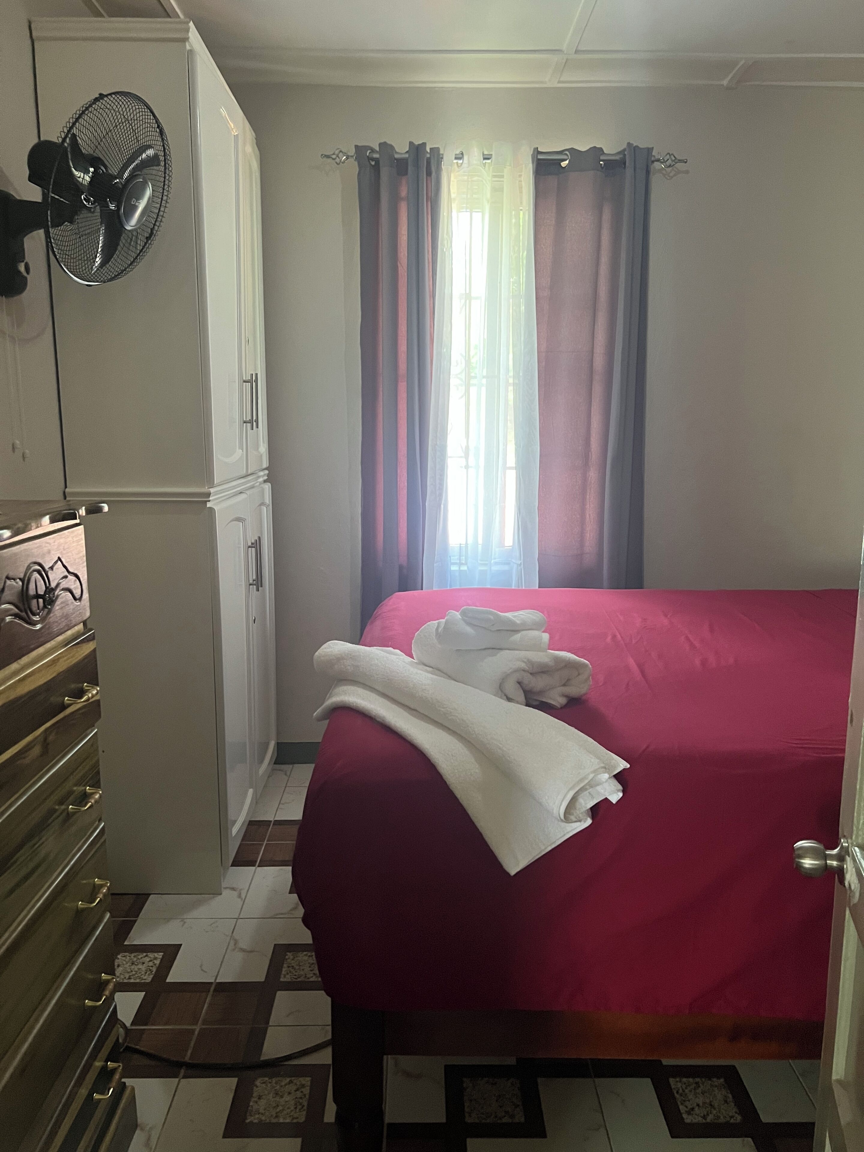 Een kamer met een bed, een ventilator, een kledingkast en een raam met gordijnen.