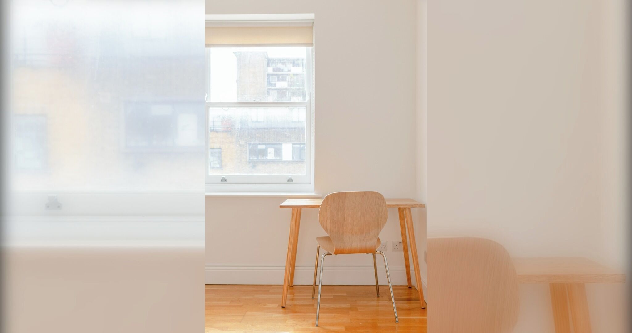 Ein Zimmer mit einem einzelnen Sessel und einem kleinen Tisch, ein Fenster mit Blick auf Gebäude und ein Holzboden.