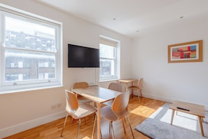 A modern living room with a wooden dining table, four chairs, a flat-screen TV mounted on the wall, and large windows offering a view of the city.
