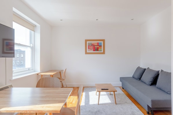 A modern living room with a sofa, a dining table, and a small coffee table. There is a window with a view of buildings outside.
