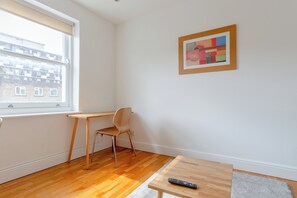 Une pièce comprenant un bureau en bois, une chaise, une petite table avec une télécommande et une fenêtre avec des stores.