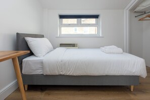 A single-bed room with a grey headboard, white bedding, a wooden bedside table, and a window with a view of buildings. - Spacious and Newly Renovated 2BD Flat Camden Town (London)