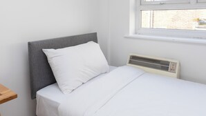 A single bed with a grey headboard and white bedding in a room with a window and a wall-mounted heater. - Spacious and Newly Renovated 2BD Flat Camden Town (London)