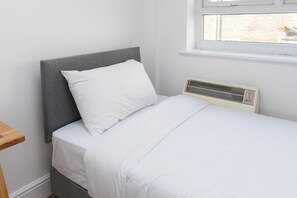 A single bed with a grey headboard and white bedding in a room with a window and a wall-mounted heater. - Spacious and Newly Renovated 2BD Flat Camden Town (London)