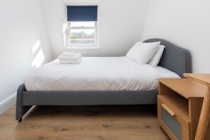 Un lit bien fait, avec du linge de lit blanc, une tête de lit grise et une table de chevet en bois dotée de tiroirs.