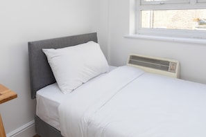 A single bed with a grey headboard and white bedding in a room with a window and a wall-mounted heater.