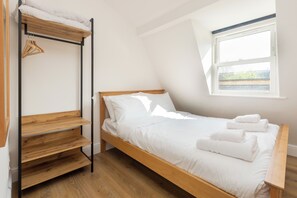 A single bed with white bedding and a wooden headboard, a wooden shelf unit with hanging clothes, and a window with a view of greenery.