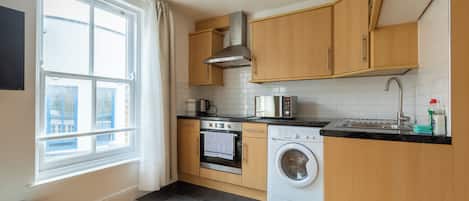 A modern kitchen with wooden cabinets, a stainless steel range hood, a built-in oven, a washing machine, and a window with curtains.