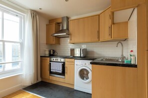 A modern kitchen with wooden cabinets, a stainless steel range hood, a built-in oven, a washing machine, and a window with curtains. - Spacious and Newly Renovated 2BD Flat Camden Town (London)