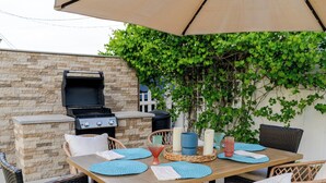 Eine Terrasse im Freien mit Tisch, Stühlen, einem Grill und einem großen Sonnenschirm.