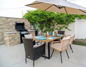 An outdoor patio with a stone wall, a grill, wicker chairs, a table with placemats, and a large umbrella. - Movie Nights & Firepit-Coastal Retreat (Jacksonville)