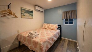 Un dormitorio pequeño y ordenado con cama, perchero, aire acondicionado, ventana con cortinas y un cuadro en la pared.