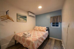 Une petite chambre à coucher, bien rangée, avec un lit, un portant à vêtements, un appareil de climatisation, une fenêtre avec des rideaux et un tableau accroché au mur.