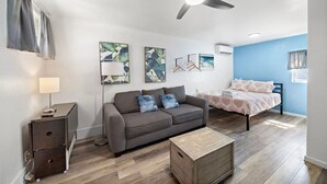 A modern bedroom with a bed, a gray sofa, a wooden nightstand, and a ceiling fan. - Oceanview Studio Near Downtown & Cruise Terminal (Galveston)