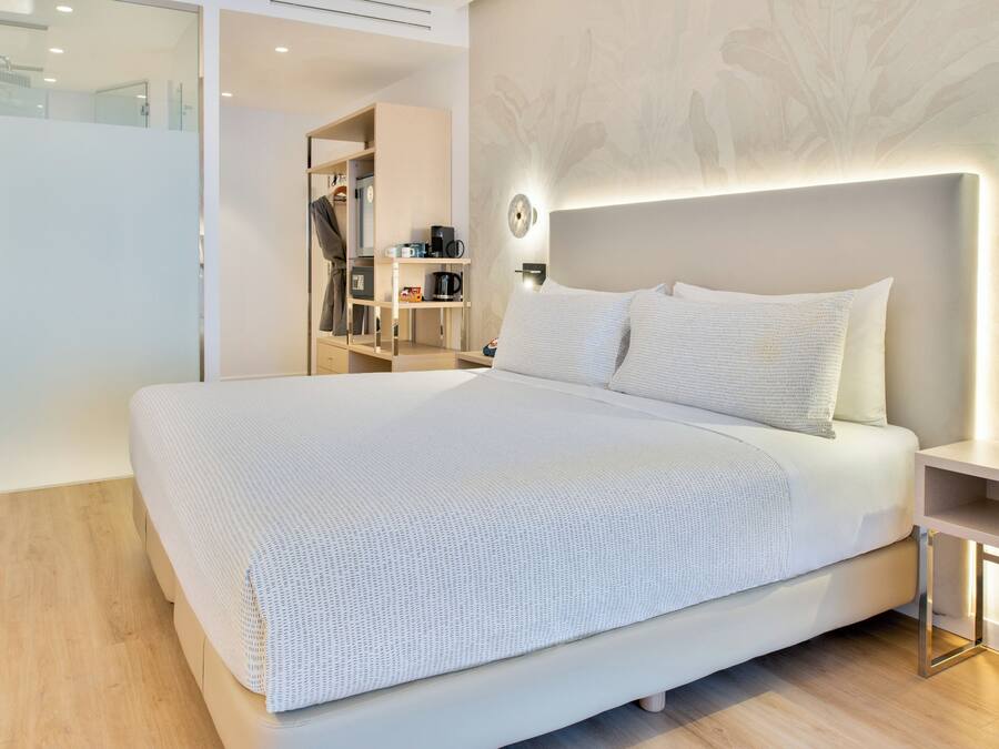 A modern hotel room with a large bed, a bedside table, a wardrobe, and a wall-mounted light fixture.