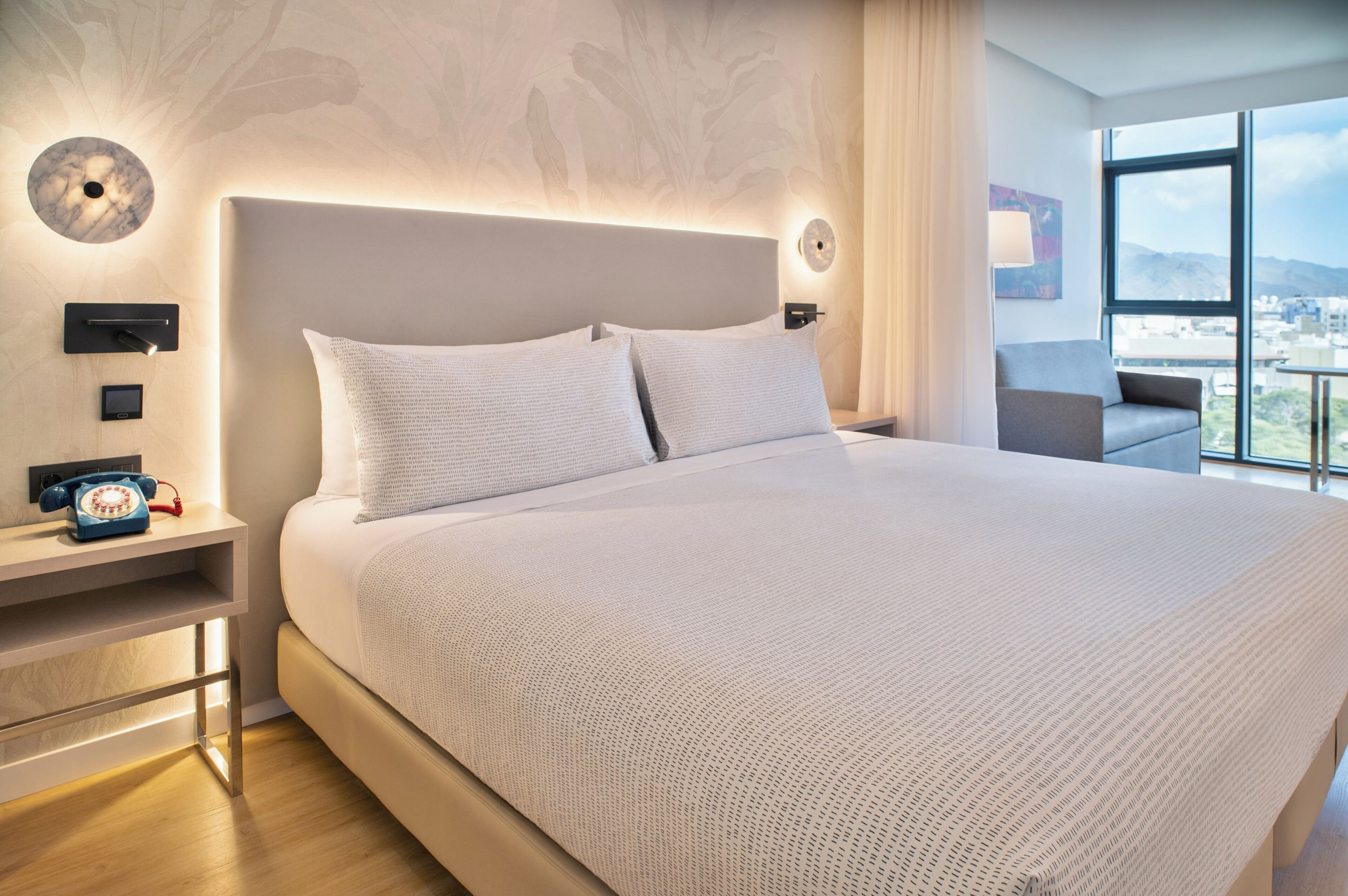 Une chambre d’hôtel moderne dotée d’un grand lit, d’une table de chevet avec un téléphone et offrant une vue sur la ville grâce à de grandes fenêtres.
