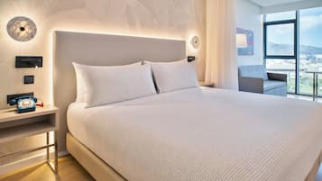 Une chambre d’hôtel moderne dotée d’un grand lit, d’une table de chevet avec un téléphone et offrant une vue sur la ville grâce à de grandes fenêtres.