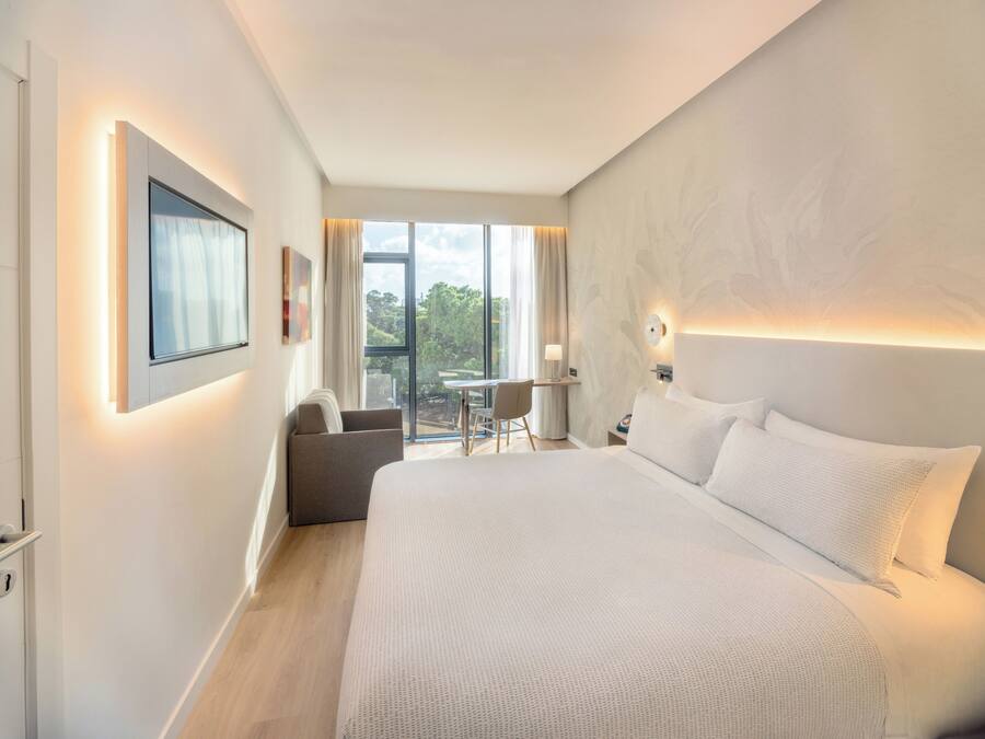 A modern hotel room with a large bed, a desk, and a balcony with a view of the city.