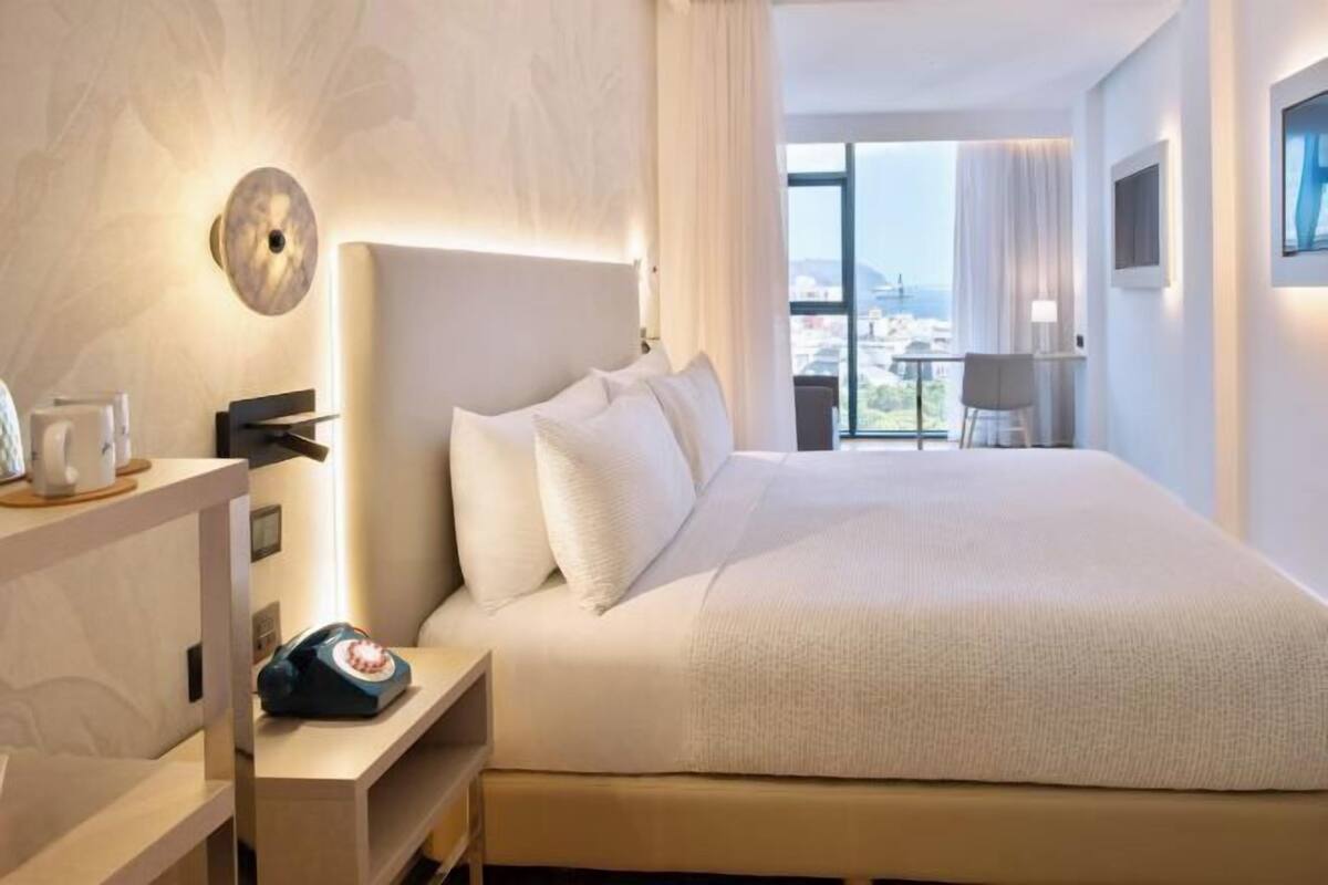 A hotel room with a large bed, a bedside table with a telephone, a wall-mounted light, and a view of the city through large windows.