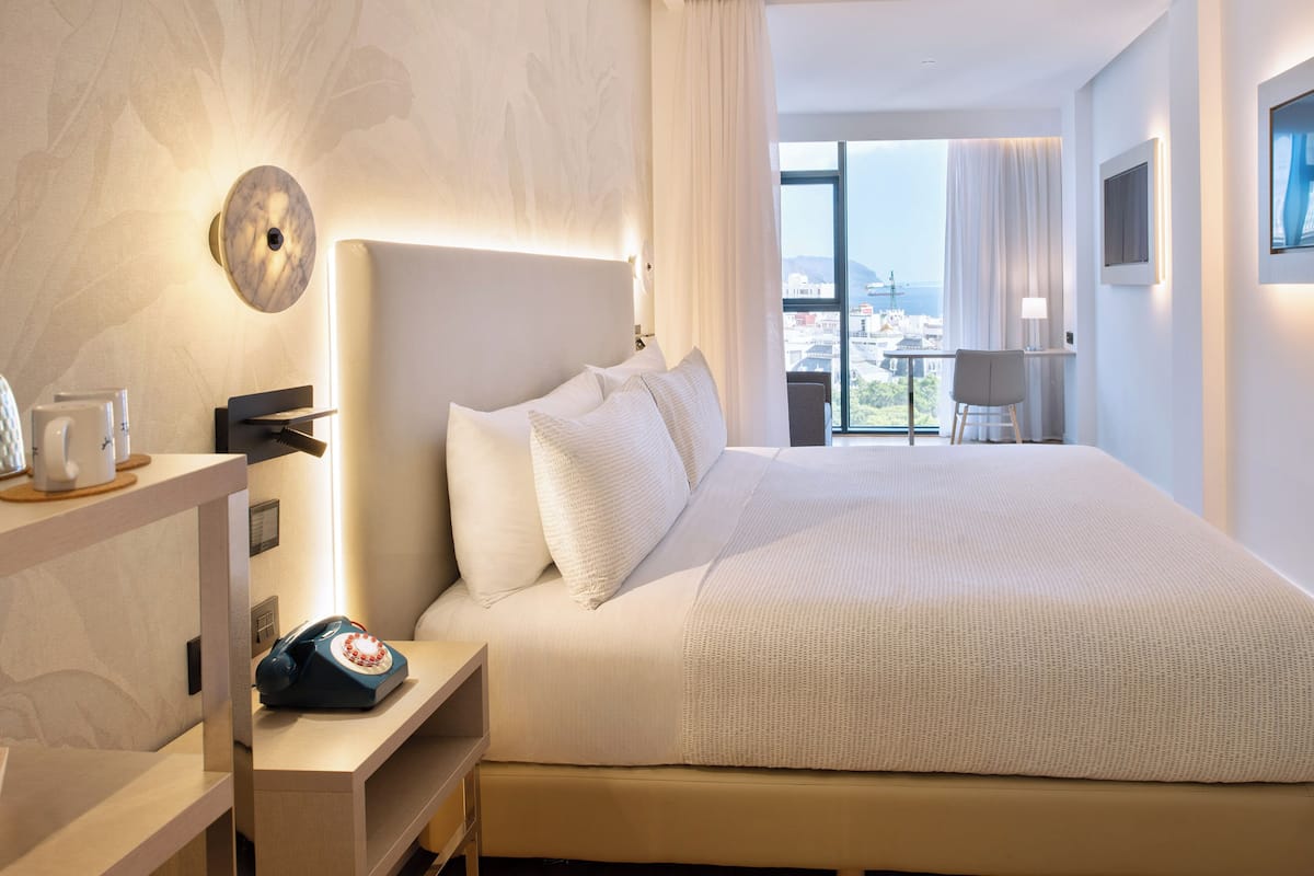 Ein modernes Hotelzimmer mit einem großen Bett, einem Nachttisch mit Telefon sowie einem Blick auf die Stadt durch große Fenster und einer städtischen Skyline im Hintergrund.