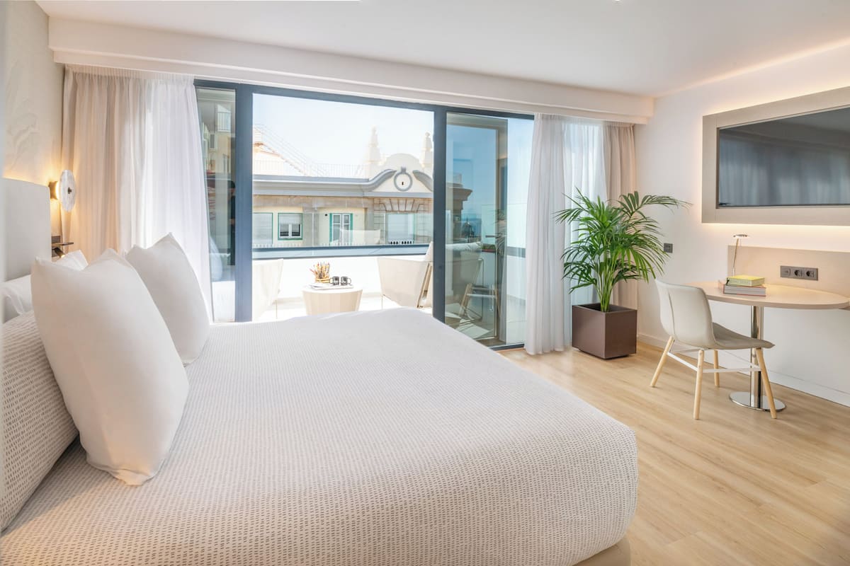 Ein modernes Hotelzimmer mit einem großen Bett, einem kleinen Tisch mit Stuhl, einer Zimmerpflanze und einem Balkon mit Blick auf Gebäude.