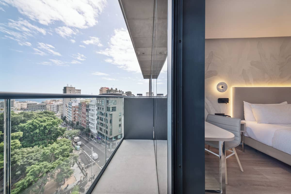 Ein modernes Hotelzimmer mit Balkon, einem Bett, einem kleinen Tisch und Blick auf die Stadtlandschaft.