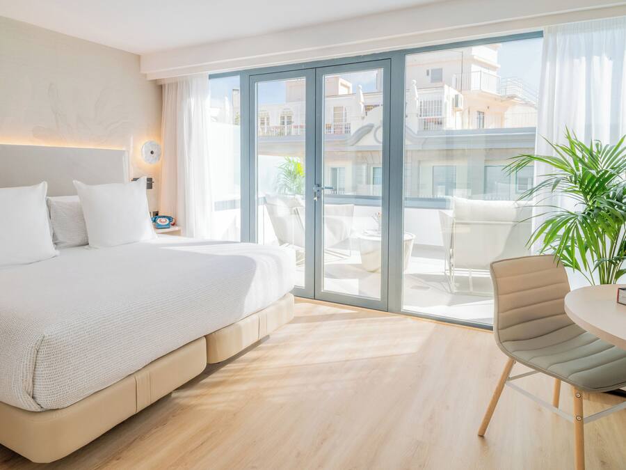 A modern hotel room with a large bed, a balcony with a view of buildings, a round table, and a chair.
