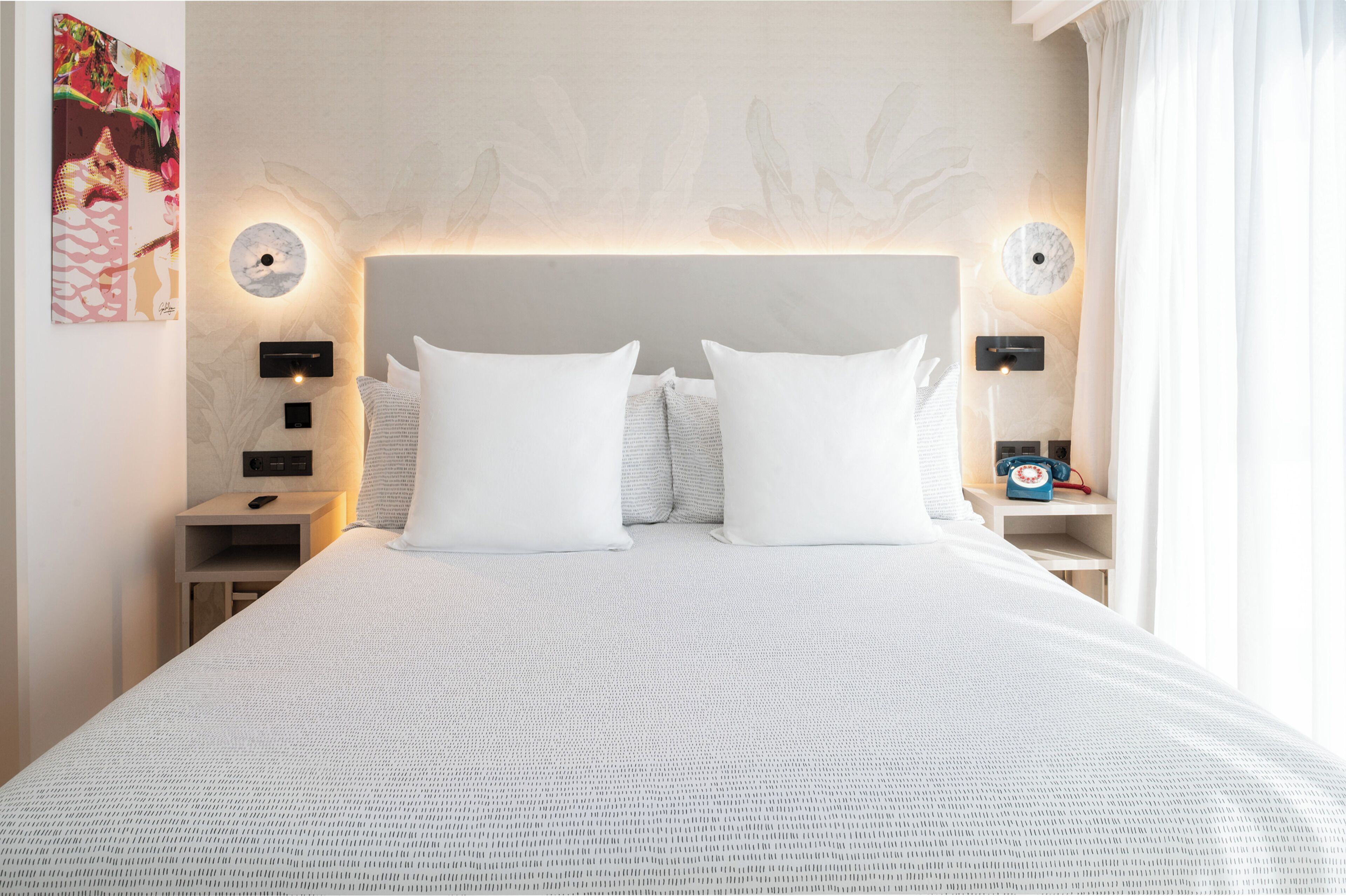 Ein ordentlich bezogenes Bett mit weißen Leinen und Kissen, zwei Nachttische mit Lampen, ein an der Wand hängendes Kunstwerk und ein Telefon auf der rechten Seite.