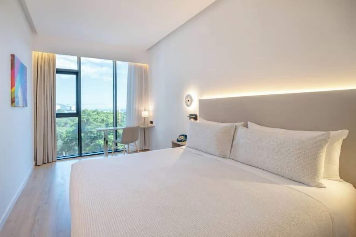 Ein modernes Hotelzimmer mit einem großen Bett, einem Schreibtisch und Blick auf Grün durch das Fenster.