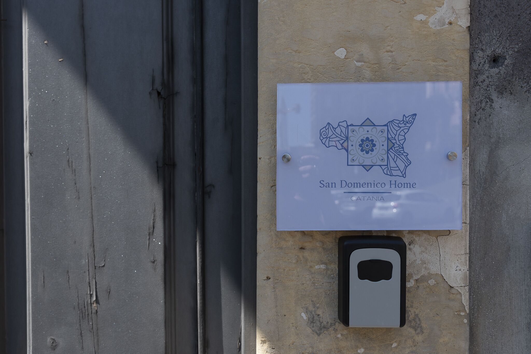 A sign with a decorative border and the text "San Domenico Home" and "Catania" on a beige wall.