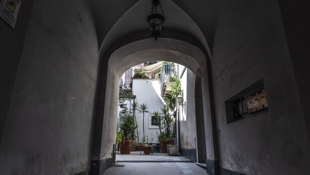 Un corredor arqueado con una lámpara colgante, que conduce a un patio con plantas en macetas y un edificio blanco al fondo.