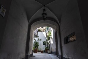 Un couloir en arc surmonté d’un luminaire suspendu, qui mène à une cour intérieure agrémentée de plantes en pot et où l’on aperçoit, au fond, un bâtiment blanc.