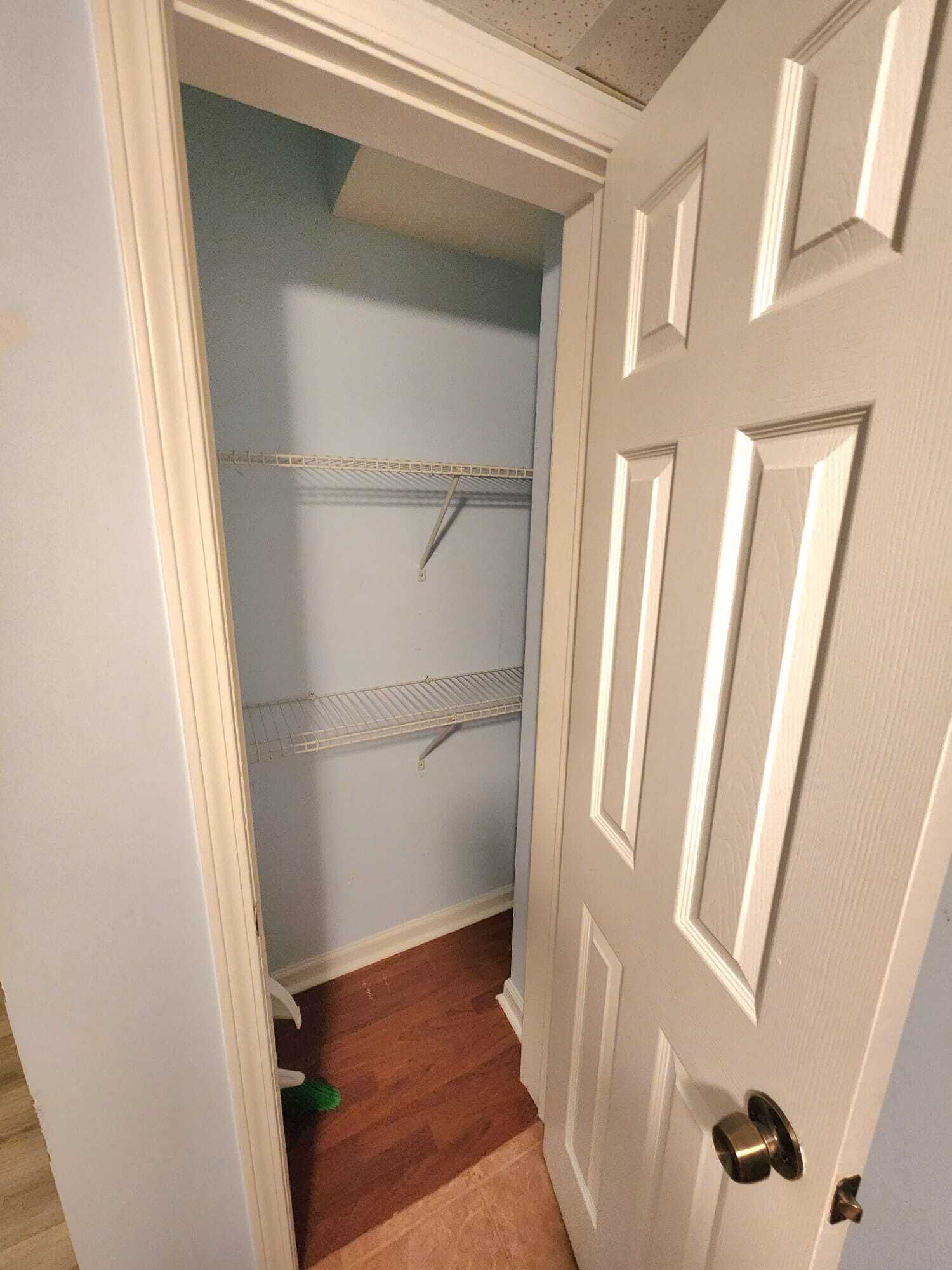 A narrow hallway with a white door on the right, a wooden floor, and two metal shelves against the wall.
