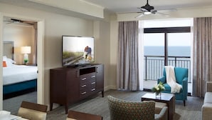 Une chambre d’hôtel avec un lit, un téléviseur posé sur une commode en bois, un balcon avec des rideaux et un coin salon comprenant un fauteuil bleu et un canapé.