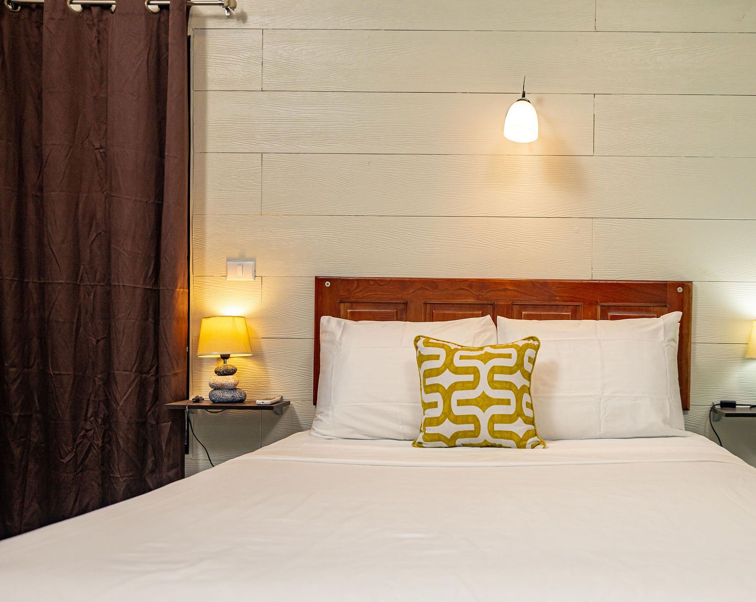 A neatly made bed with white linens, a wooden headboard, and a patterned pillow. There is a bedside lamp on a wooden nightstand, and a wall-mounted light fixture above the bed.
