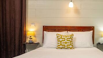A neatly made bed with white linens, a wooden headboard, and a patterned pillow. There is a bedside lamp on a wooden nightstand, and a wall-mounted light fixture above the bed.