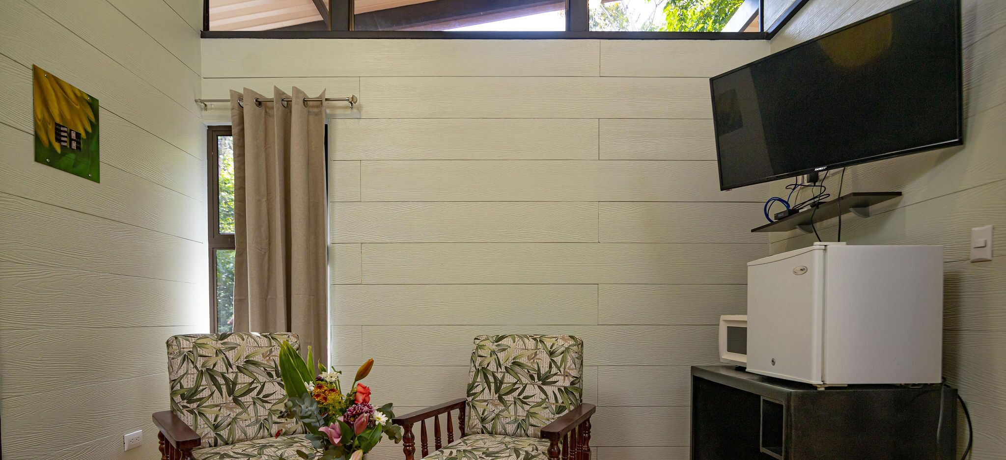 A room with two chairs, a small table with a floral arrangement, a television mounted on the wall, and a microwave on a counter.