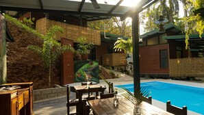 A covered outdoor area with a pool, wooden furniture, and a painting of a frog. - Riverside Bungalows (Quepos)