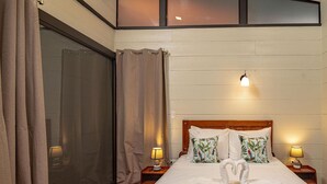 A bedroom with a large bed, two nightstands, a window with blinds, and a curtain. - Riverside Bungalows (Quepos)