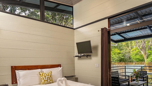 A bedroom with a bed, a TV mounted on the wall, and a view of an outdoor area with a table and chairs. - Riverside Bungalows (Quepos)