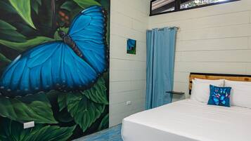 Un dormitorio con un gran mural de una mariposa azul, una cama con ropa de cama blanca y almohadas azules, y una ventana con cortina azul.