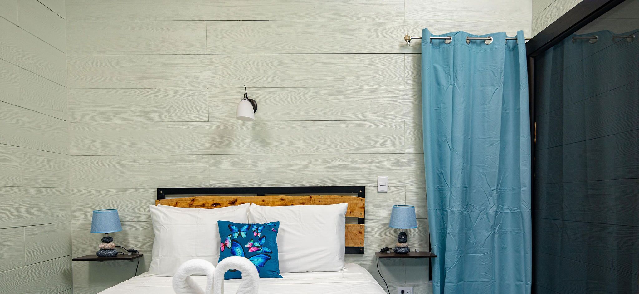 Habitación de hotel con cabecera de madera, ropa de cama blanca, almohada con forma de mariposa azul, dos lámparas de noche y cortina azul.