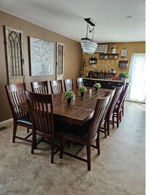 A dining area with a long wooden table surrounded by chairs, a chandelier, and a bar area with shelves and bottles.