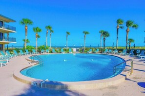 Une piscine dont la profondeur maximale est indiquée à 7 pieds, entourée de chaises longues et de palmiers, avec un bâtiment situé sur l’un des côtés.