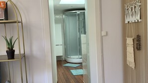 Un couloir doté d’un plancher en bois, d’une cabine de douche en verre et d’une porte blanche donnant sur une autre pièce.