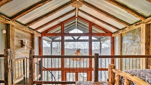 A rustic cabin interior with a wooden floor, a stone wall, and a large window offering a view of the outdoors.