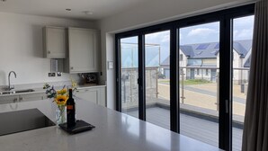 A modern kitchen with a large island, a built-in oven, and a view of a residential area through large sliding glass doors. - Galwad y Mor (Haverfordwest)