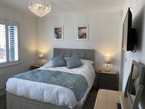 A neatly arranged bedroom with a large bed, two bedside tables with lamps, a television on the wall, and two framed pictures above the bed. - Galwad y Mor (Haverfordwest)