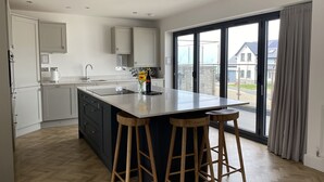 A modern kitchen with a large island, wooden stools, and a view of houses through large windows. - Galwad y Mor (Haverfordwest)